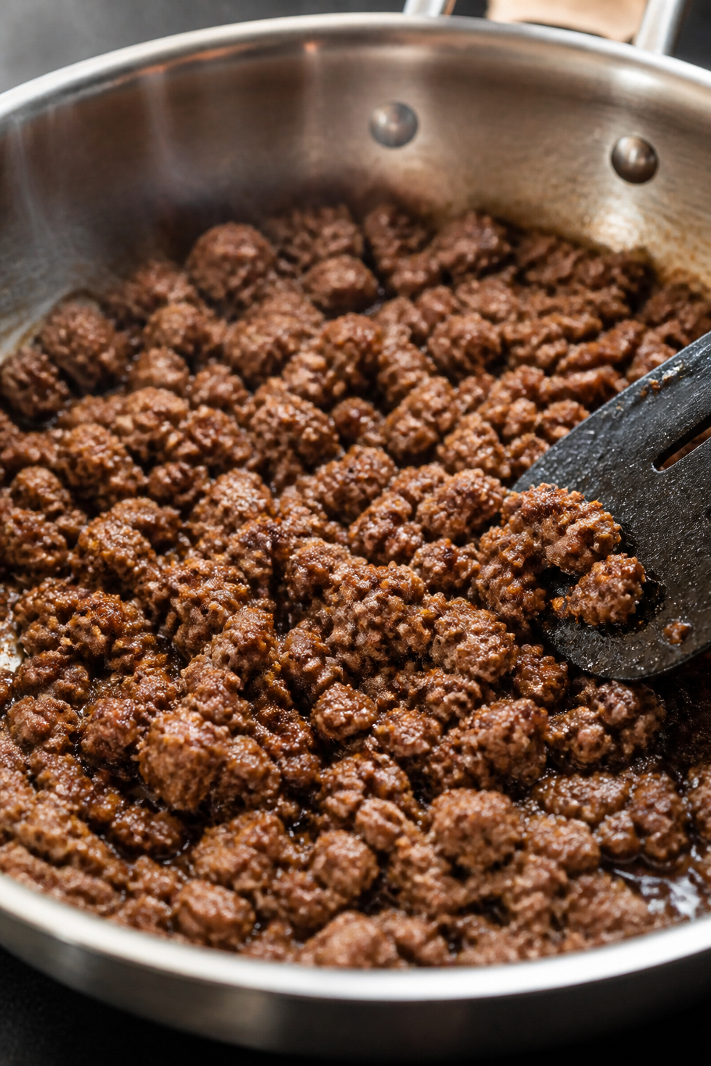 Cooking process, close-up: Crumbled lean ground beef sizzling in a wide stainless skillet, browned a